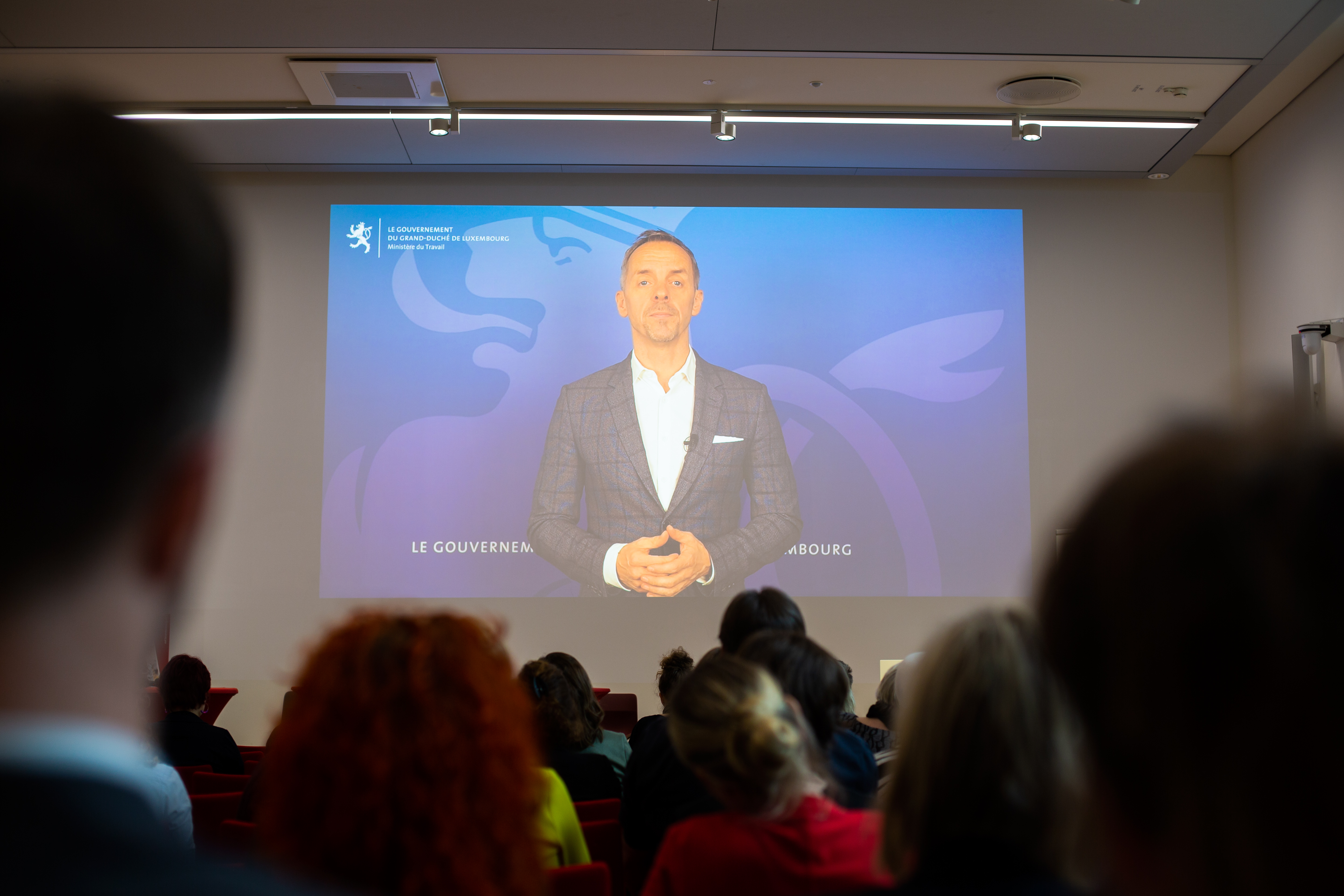 Bild: Georges Mischo, Minister für Arbeit in Luxemburg, ist auf einer Leinwand zu sehen; über die Köpfe des Publikms hinwegfotografiert.