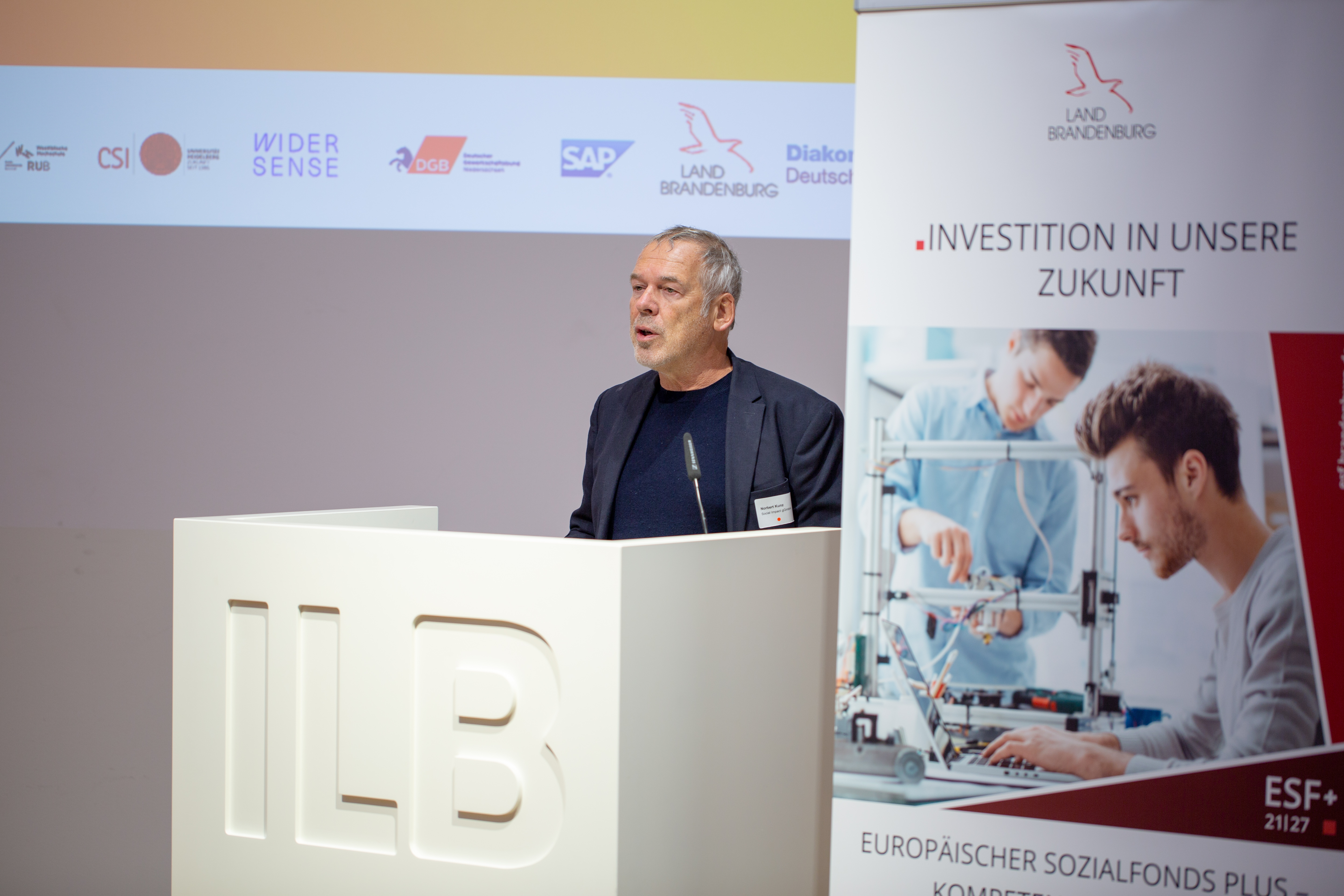 Bild: Norbert Kunz, Social Impact-Geschäftsführer, steht am Rednerpult; daneben ein ESF+-Roll-up auf dem 'Investition in unsere Zukunft' steht.