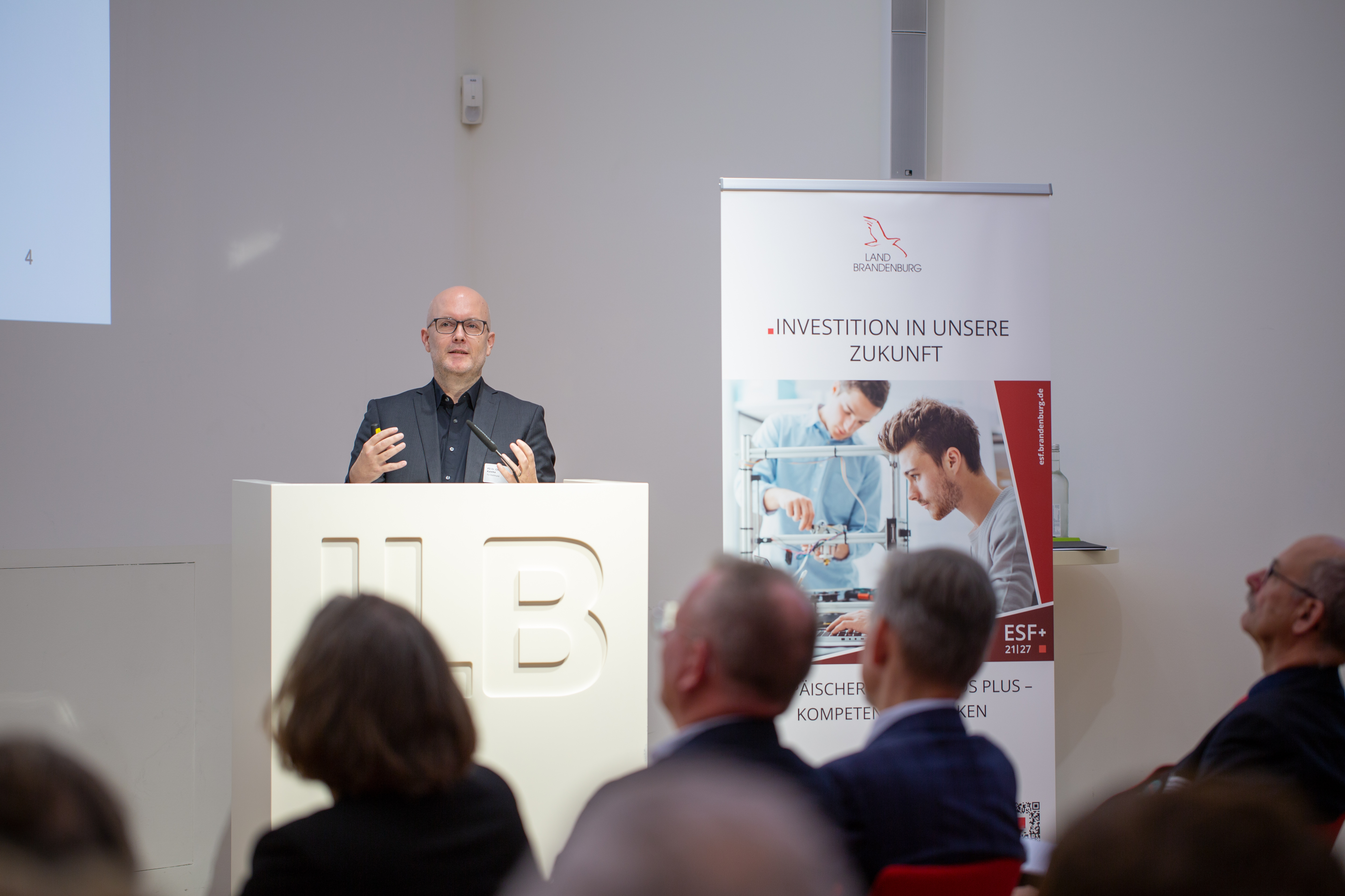 Bild: Dr. Christoph Kaletkavon der Technischen Universität Dortmund steht am ILB-Rednerpult; über die Köpfe des Publikums hinwegfotografiert.