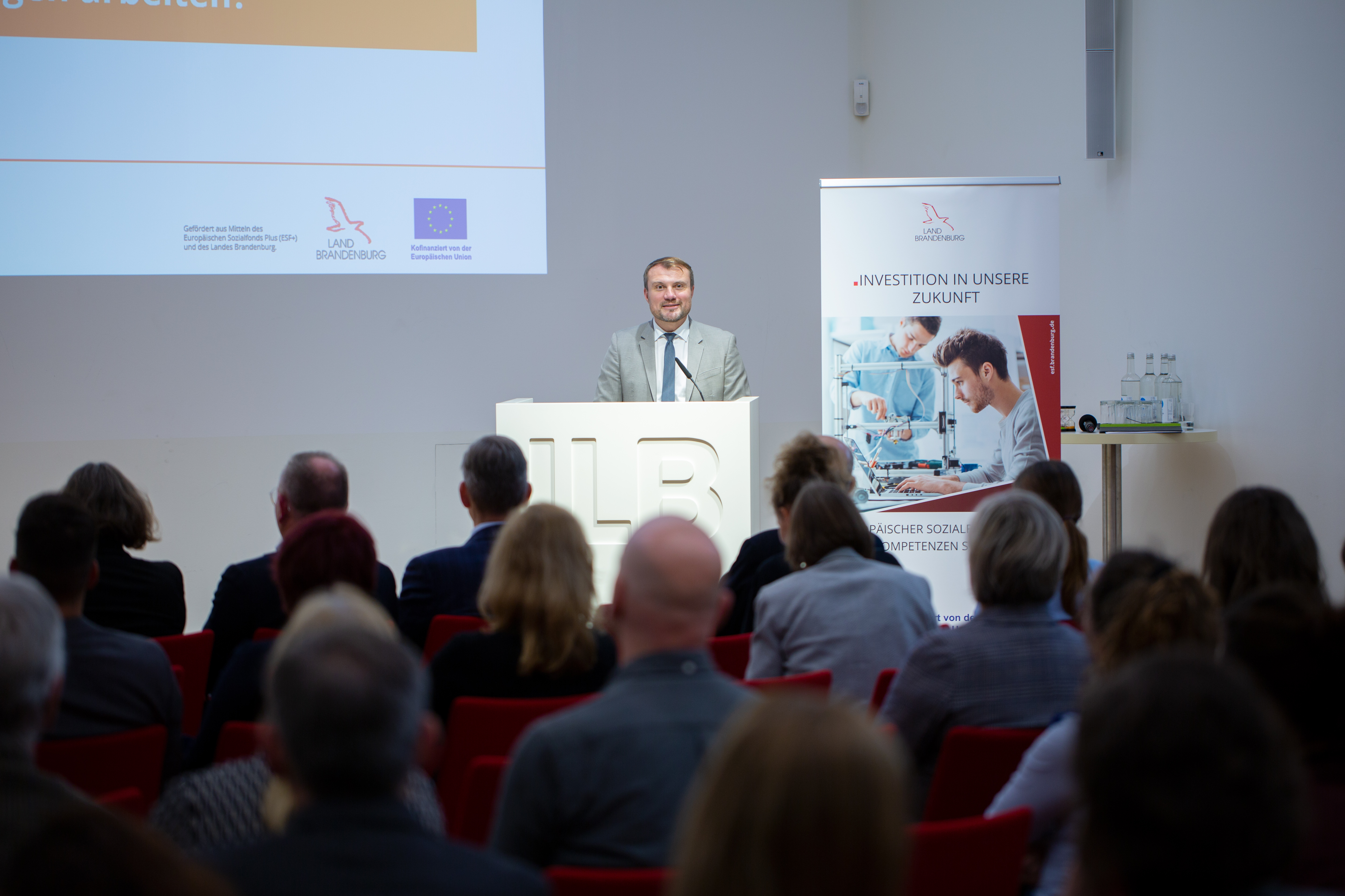 Bild: Brandenburgs Wirtschaftsminister Daniel Keller am Rednerpult; über die Köpfe des Publikums hinweg fotografiert.