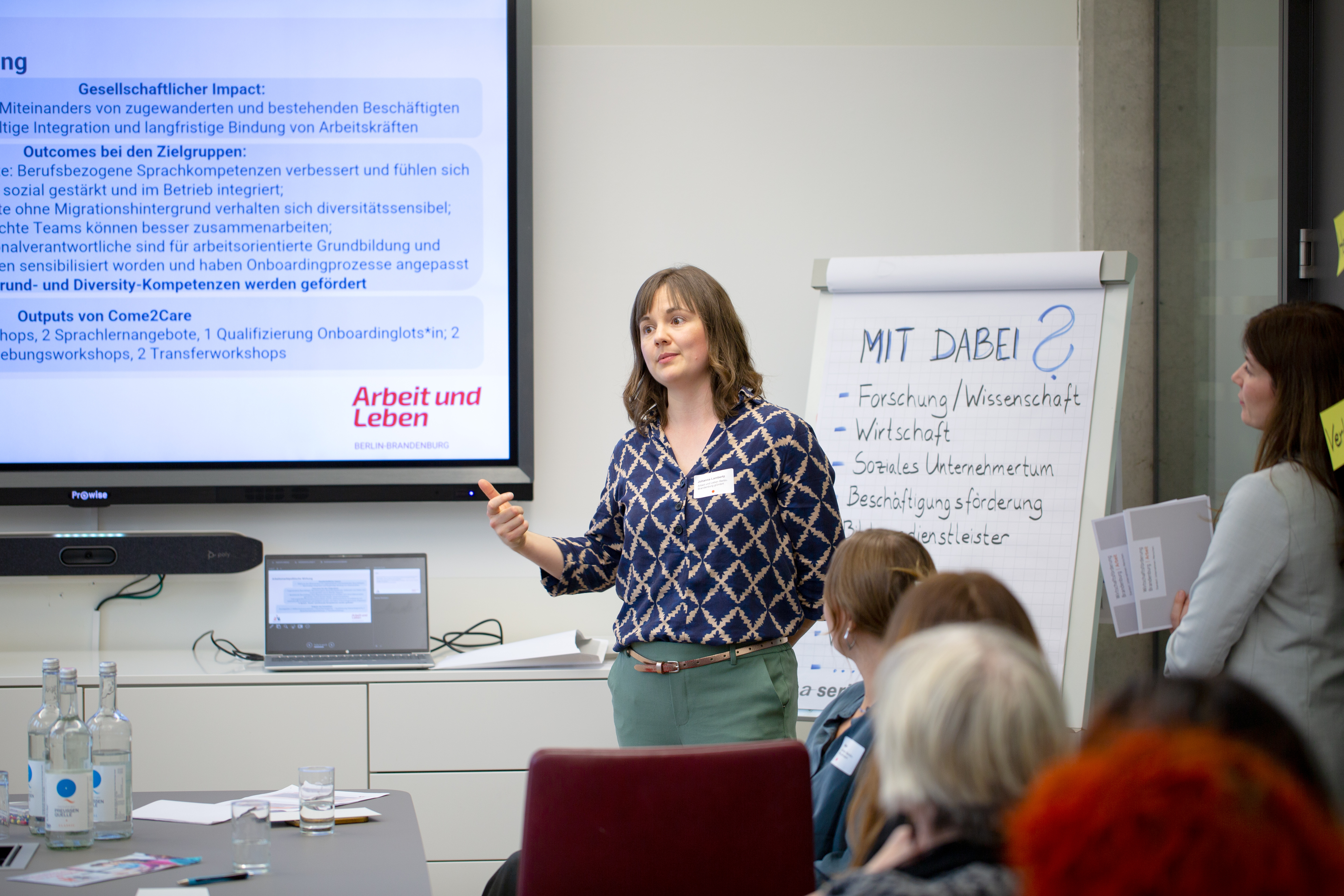 Bild: Teilnehmerin eines Workshops neben einem großen Bildschirm, rechts neben ihr ein Flipchart, im Vordergrund die Köpfe anderer Teilnehmenden.
