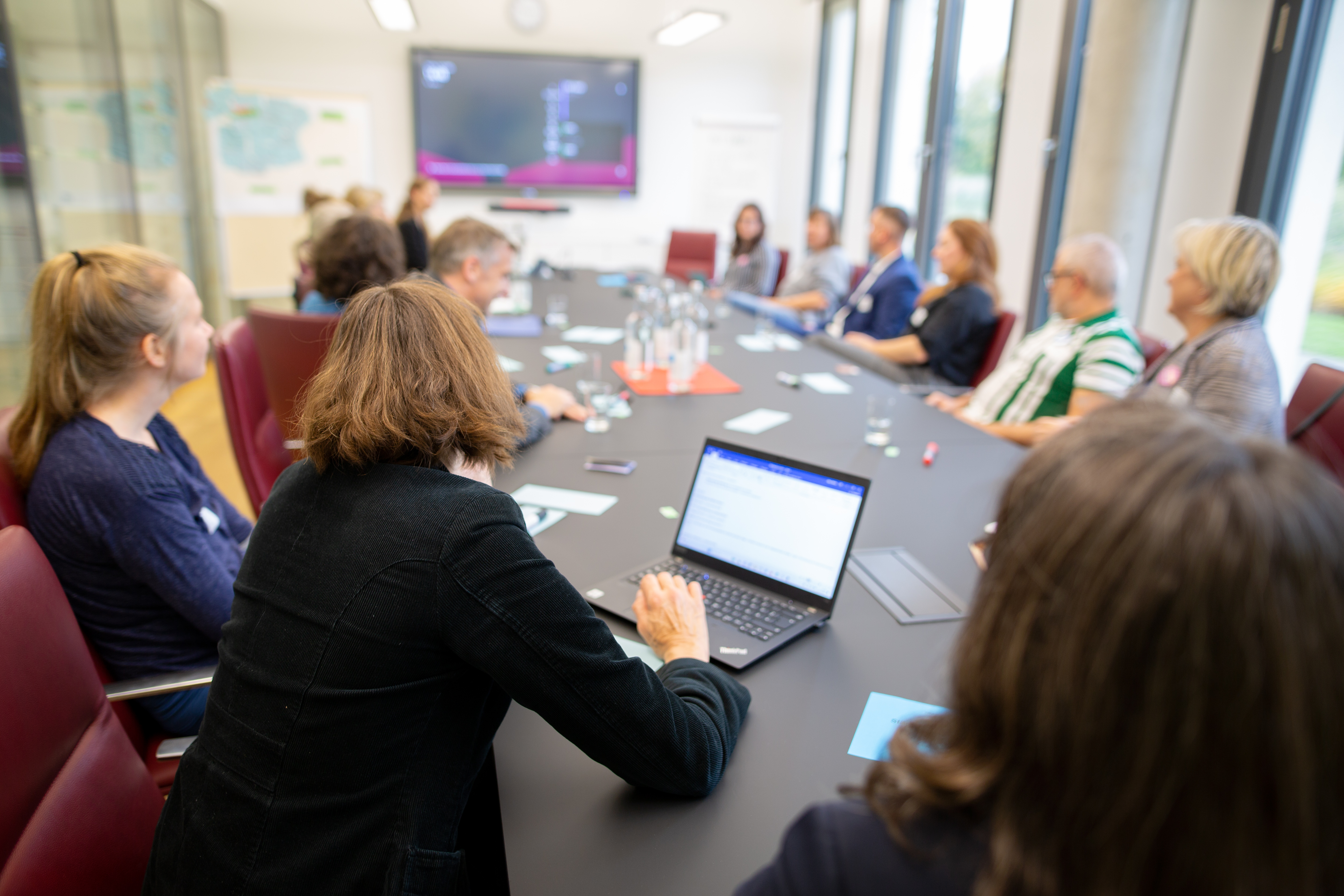 Bild: Langer Tisch mit rechts und links sitzenden Workshop-Teilnehmenden. Auf dem Tisch liegen Stifte und Zettel, im Hintergrund ein Flipchart und eine Metaplanwand.