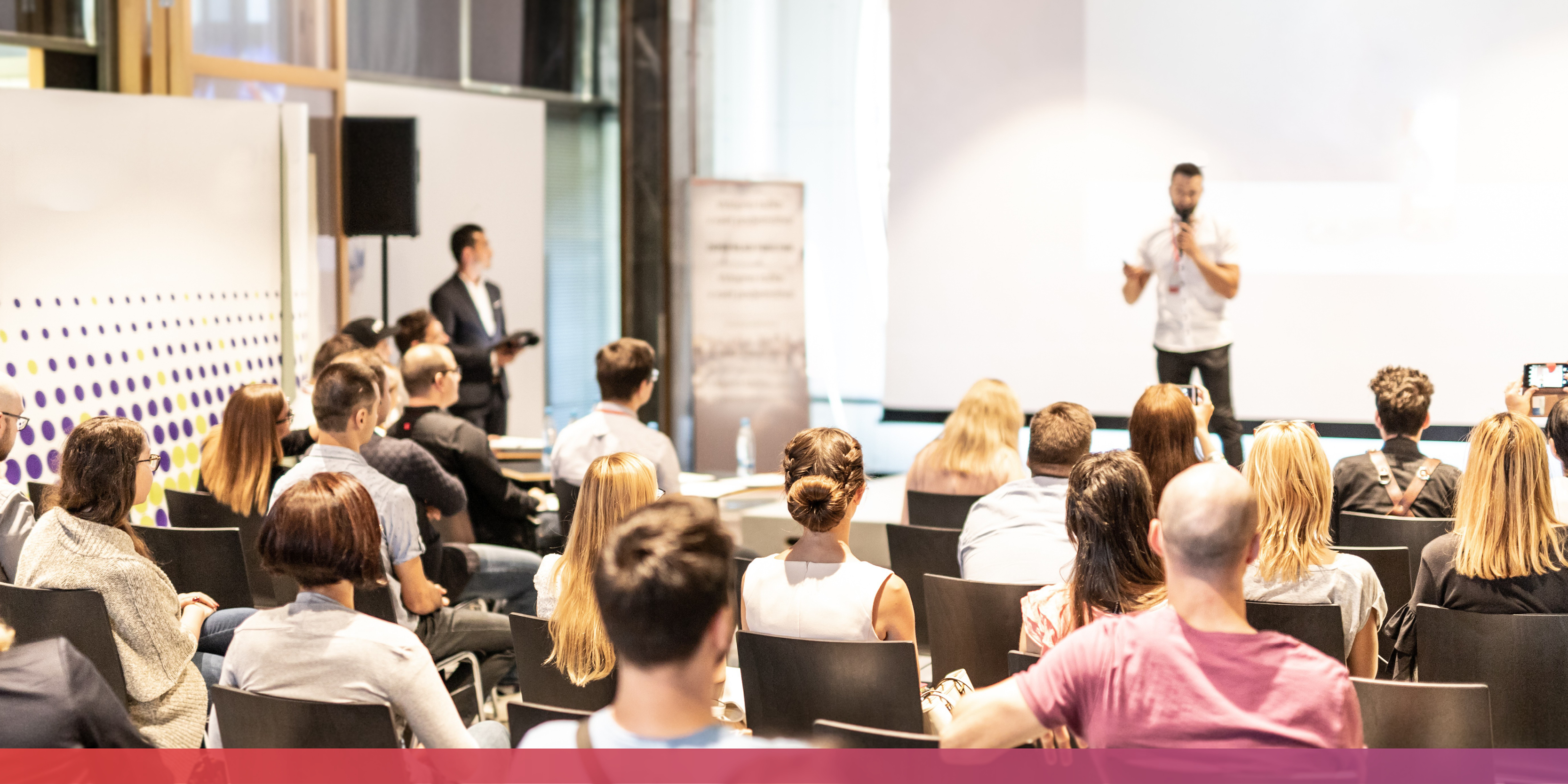Vollbesetzte Stuhlreihen und unscharf im Hintergrund erkennbar ein Mensch mit Mikrofon in der Hand, der etwas präsentiert.