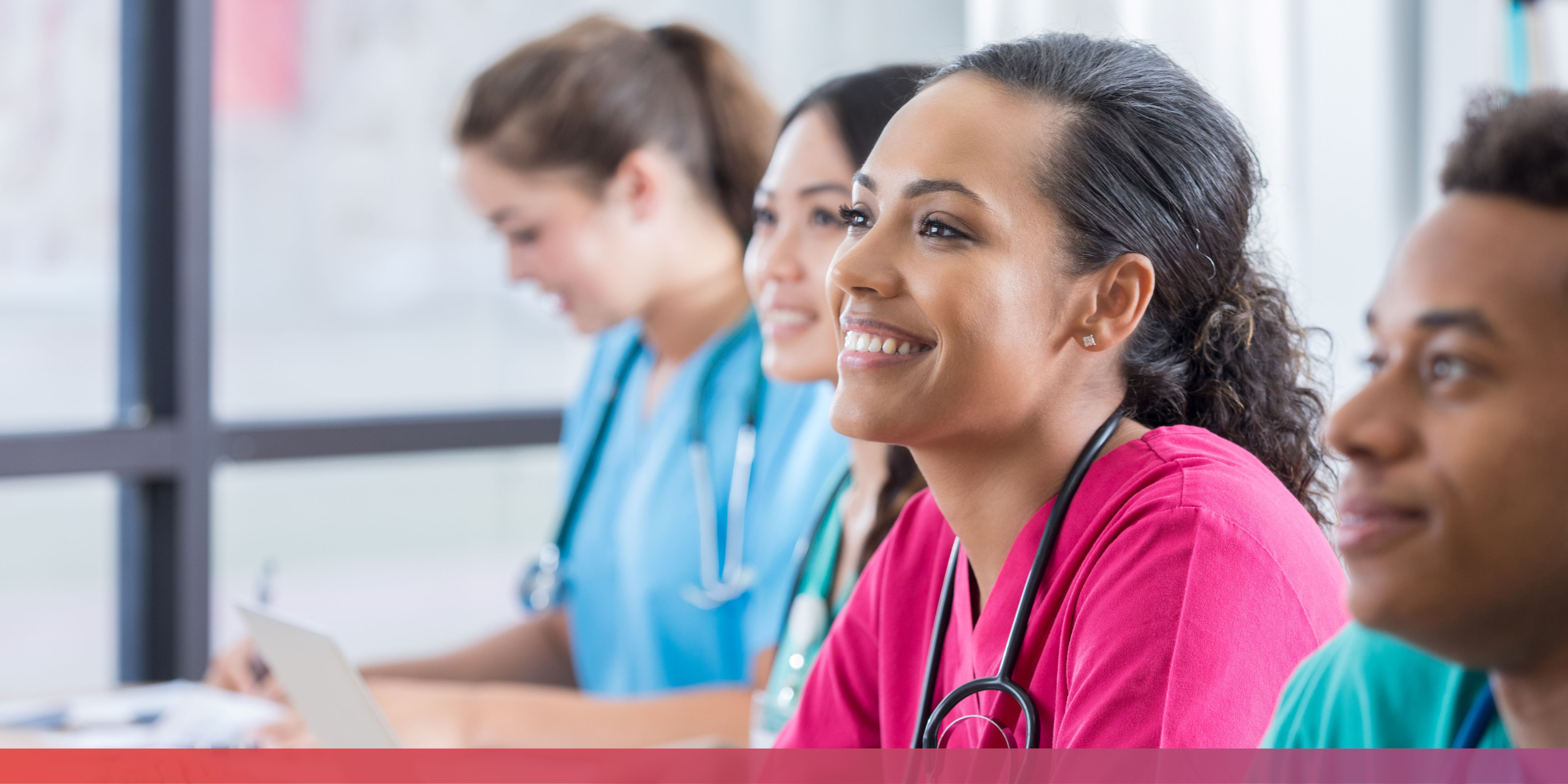 Mehrere Menschen unterschiedlicher Nationalitäten in Pflegekleidung (teils mit Stethoskop um den Hals) sitzen nebeneinander und schauen einem Referierenden zu.