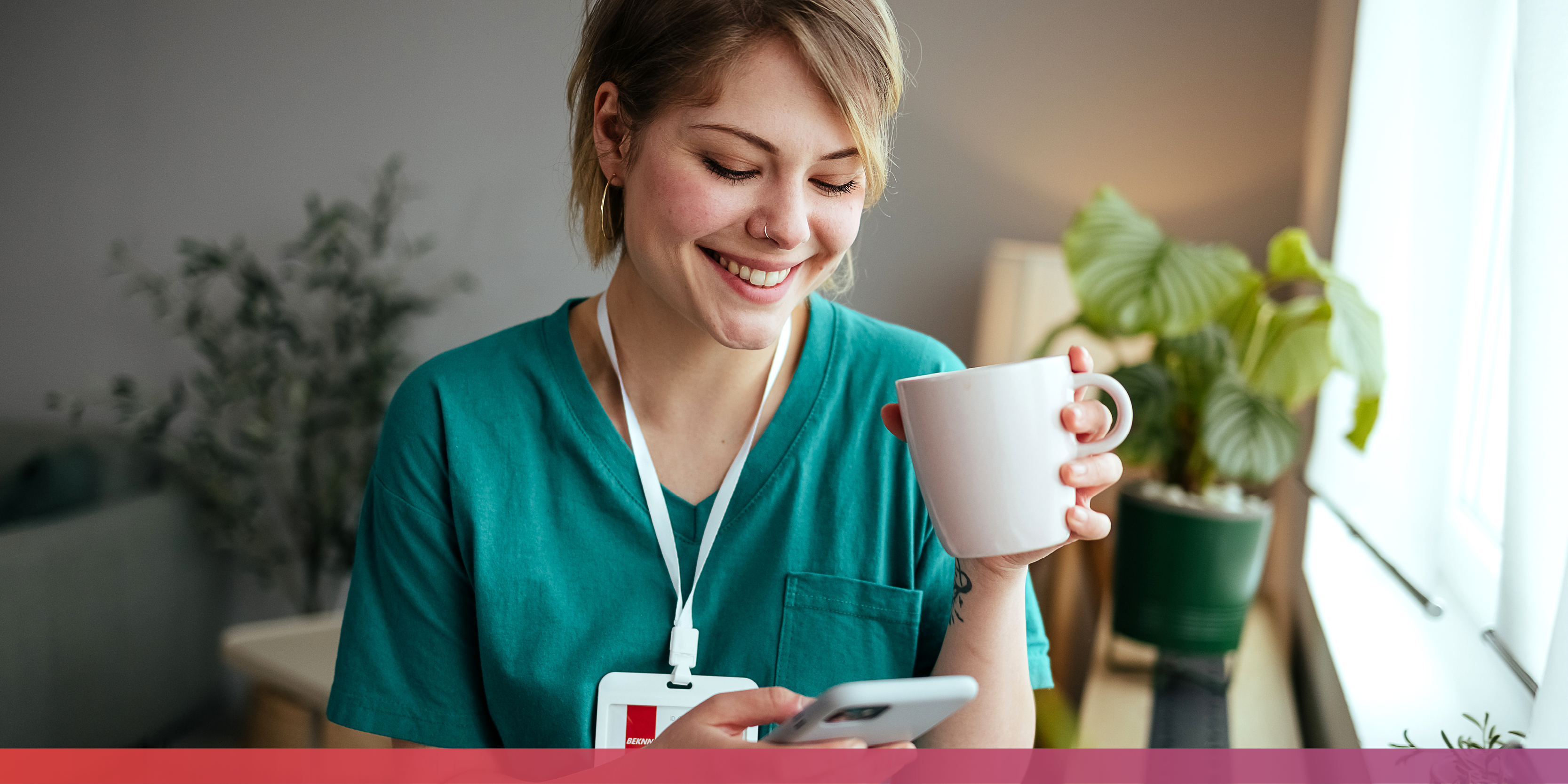 Junge, lächelnde Frau in grüner Pflegekleidung mit Kaffeetasse in der linken und Handy in der rechten Hand.