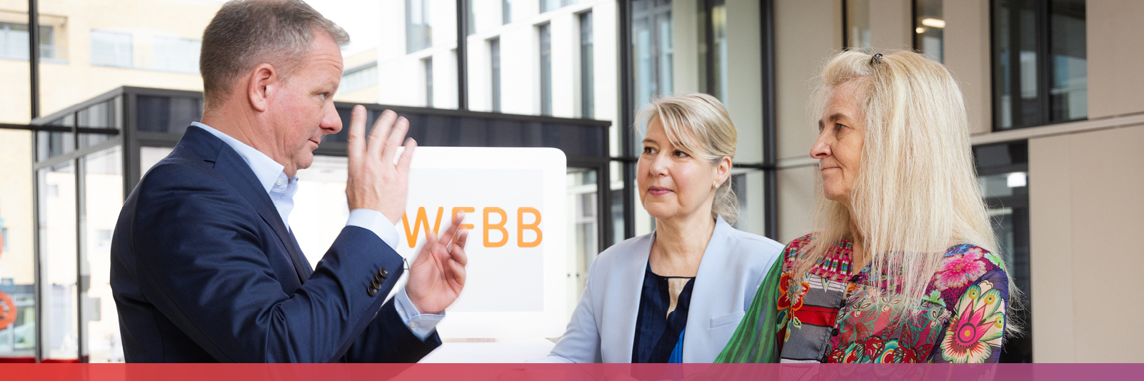 Ein Mann im Anzug steht vor zwei Frauen, die rechts von ihm stehen und berät sie gestikulierend.