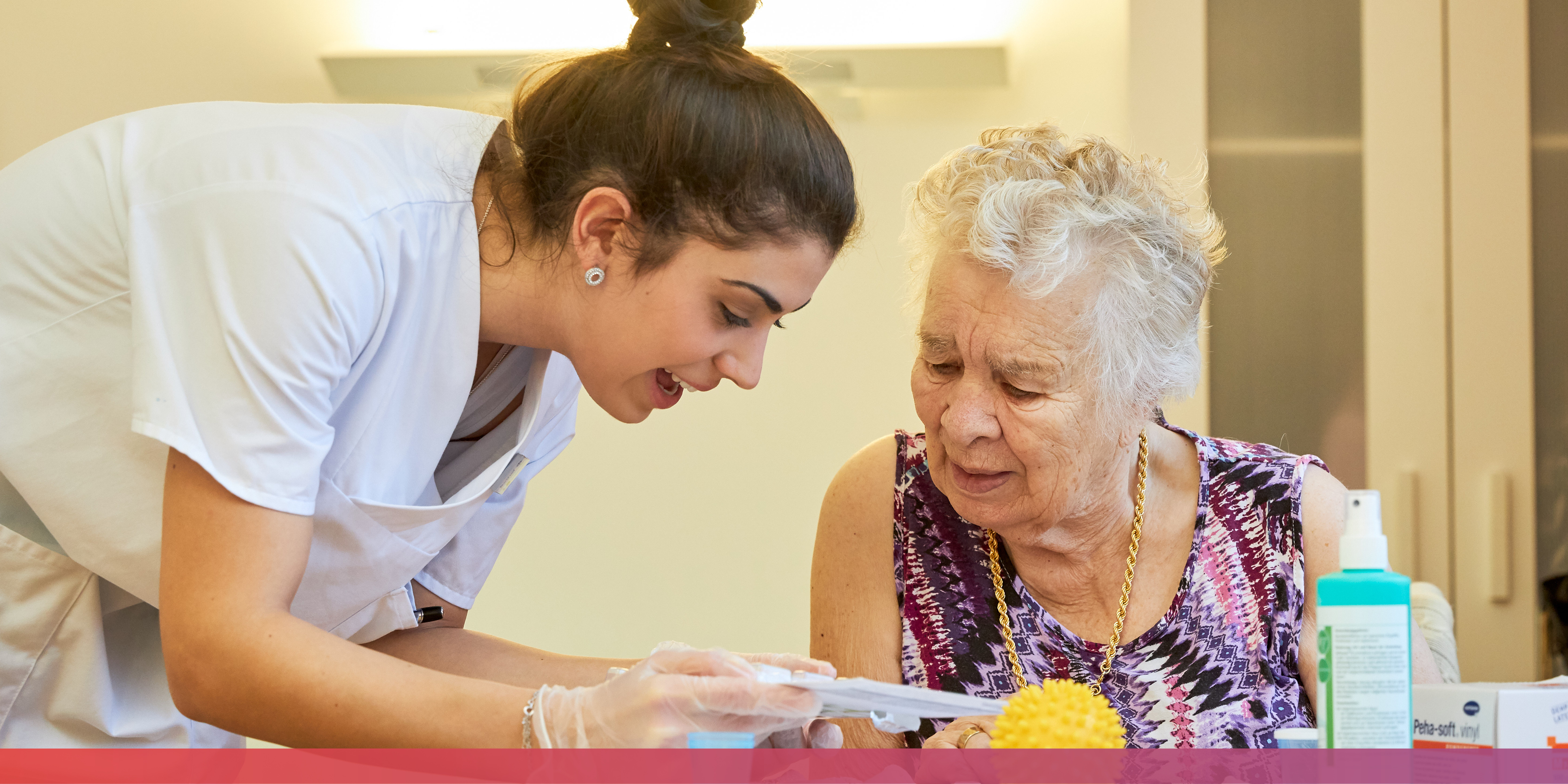 Eine junge Pflegekraft beugt sich zu einer älteren Dame herunter und liest ihr etwas vor.
