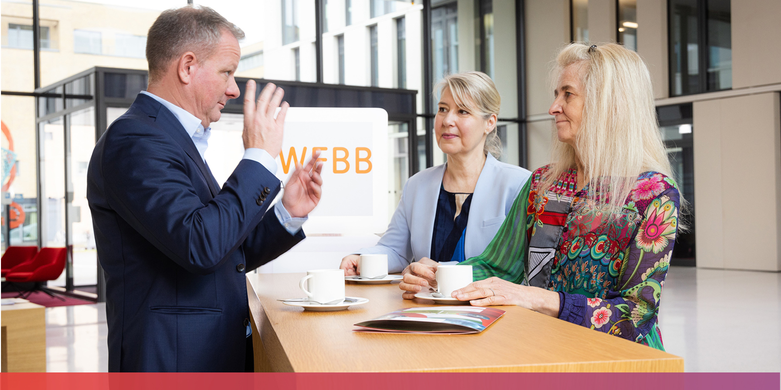 Bild: Ein Mann im Anzug steht vor zwei Frauen, die rechts von ihm stehen und berät sie gestikulierend.