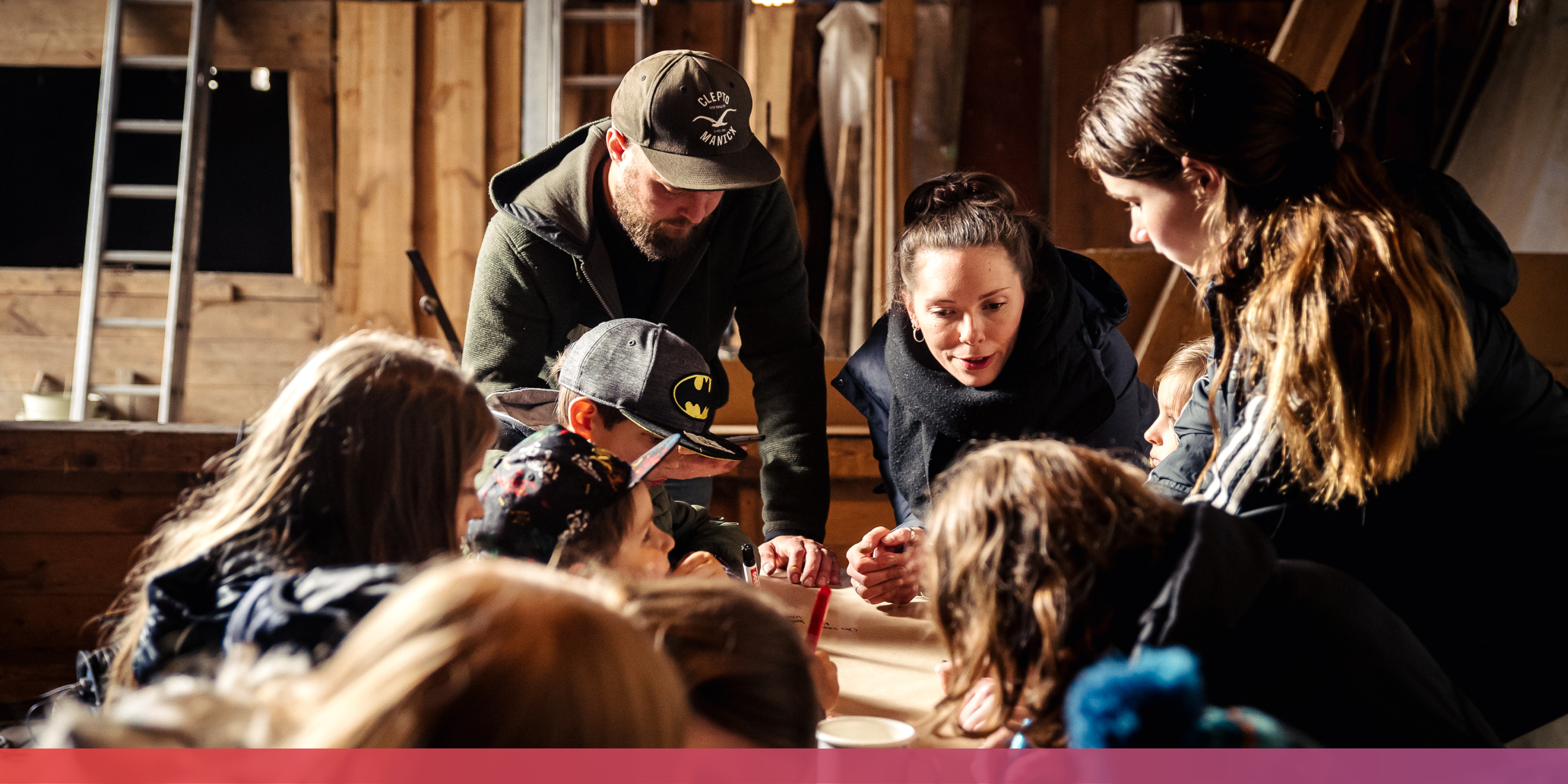 Junge Erwachsene und Kinder stehen gebückt bzw. sitzen um einen Tisch herum und unterhaltzen sich; im Hintergrund eine Werkstatt oder eine Scheune.