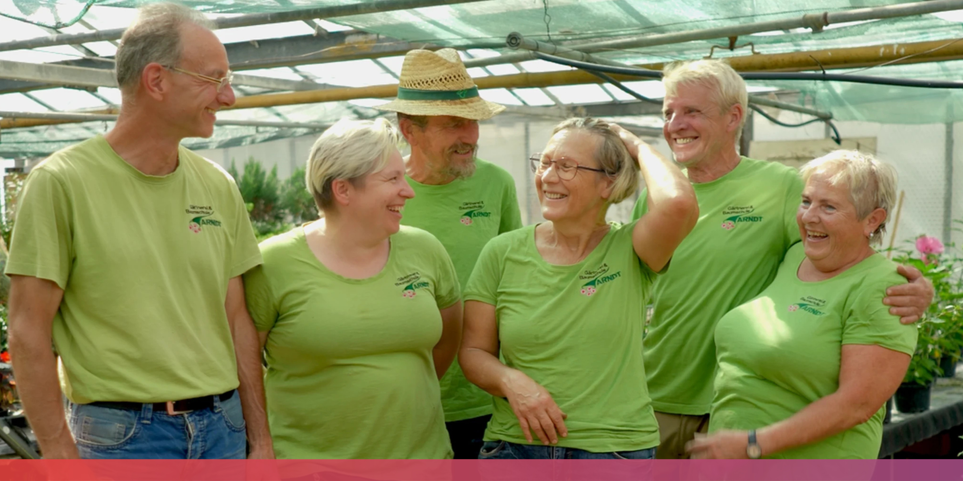 Mehrere ältere Menschen in grünen T-Shirts, die sich teils umarmen und alle lächeln; im Hintergrund ein Gewächshaus. 