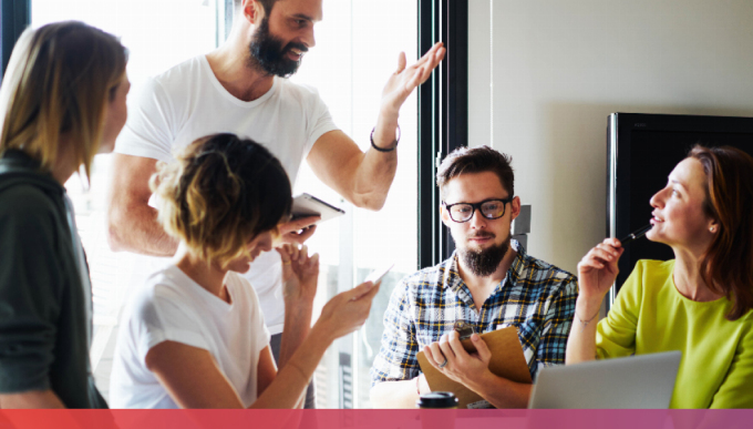 Mehrere Menschen sitzen an einem Tisch in einer Art Brainstorming und unterhalten sich angeregt, teils haben sie Tablets, Handys oder ein Klemmbrett in der Hand.