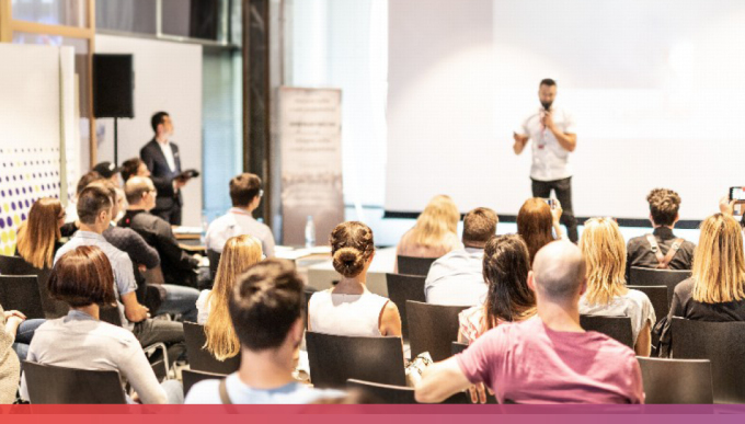 © AdobeStock: kasto Vollbesetzte Stuhlreihen und unscharf im Hintergrund erkennbar ein Mensch mit Mikrofon in der Hand, der etwas präsentiert.